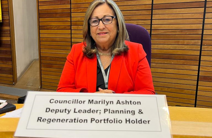 Cllr Marilyn Ashton sat in the old Council Chamber dressed in a red blazer looking at the camera and smiling. In front of her is her name plaque which reads 'Councillor Marilyn Ashton: Deputy Leader, Planning & Regeneration Portfolio Holder