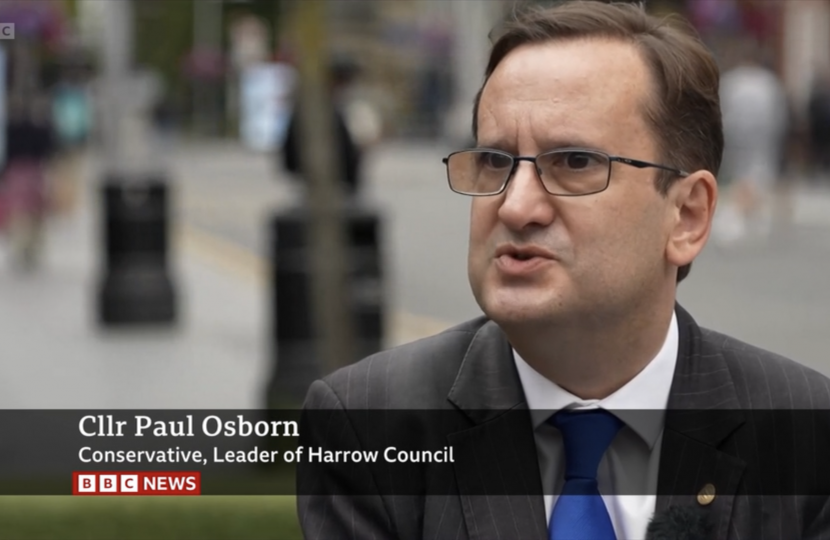 Paul Osborn in Harrow Town Centre wearing a suit, white shirt and a blue tie. In the background are people shopping.