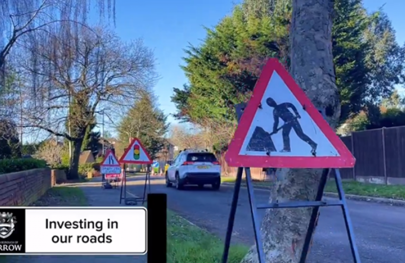 A photo showing a warning sign for road works. In the background are workmen working on a road. There are trees and houses either side of the road.